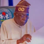 President Bola Tinubu seated at a desk reviewing papers, wearing cream traditional attire and a patterned cap.