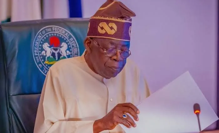 President Bola Tinubu seated at a desk reviewing papers, wearing cream traditional attire and a patterned cap.