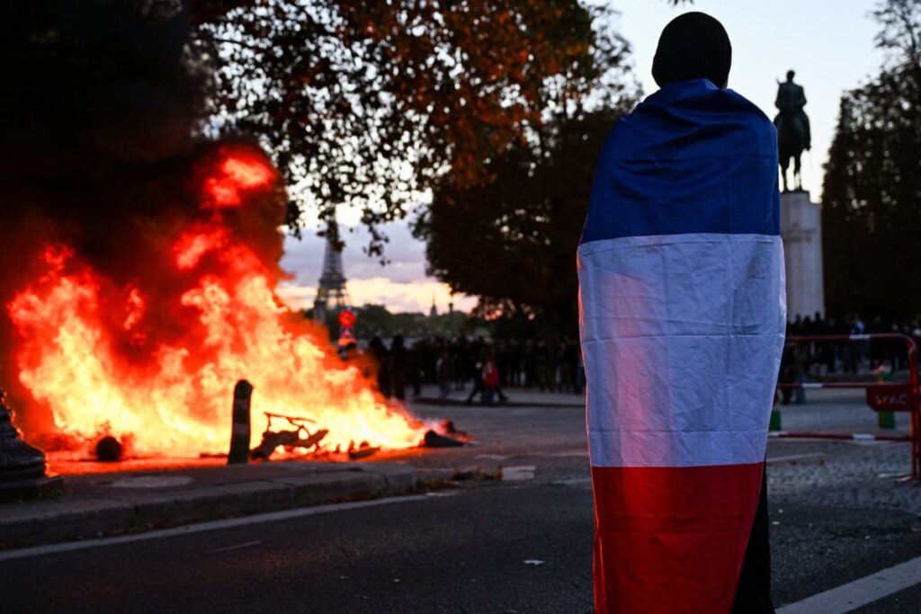 A protester draped in the French flag stands near a fire during a cost-of-living demonstration in Paris on 17 October 2026
