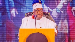 George Akume speaking at a lectern in an archive photograph, wearing a white cap and traditional attire.