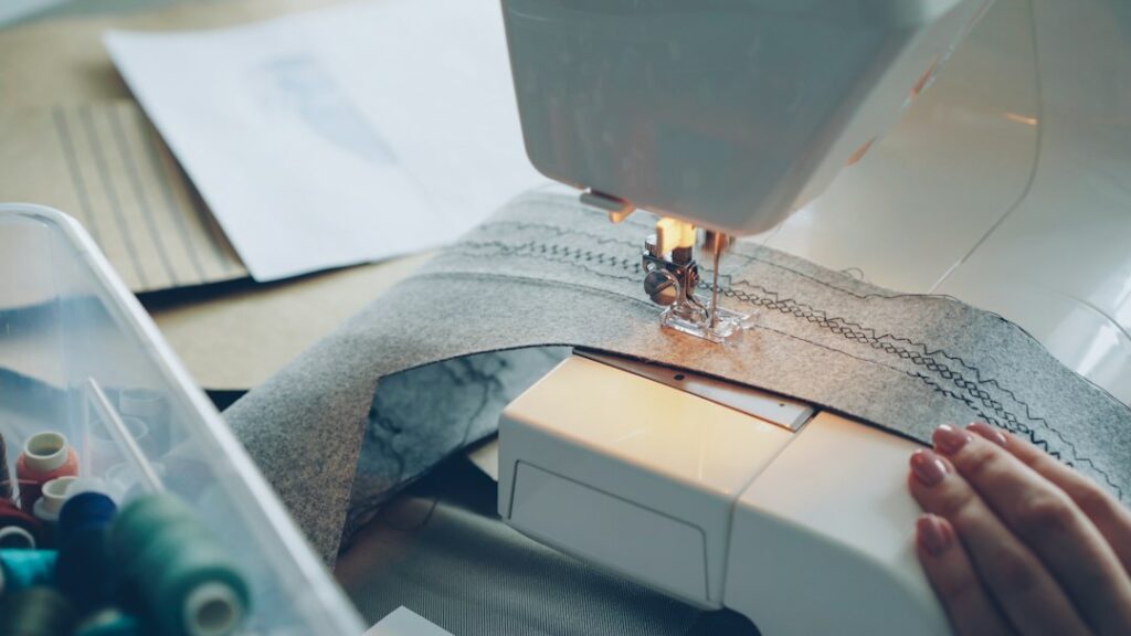 Hands guiding fabric through a sewing machine, illustrating Zimbabwe women farmers’ garment project